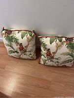 Two decorative cushions with cream colored background and illustrations of monkeys in red vests climbing leafy tree branches, standing against a white wall on wooden floor.