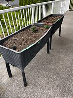 Two large black elevated planters with woven rattan-style texture filled with soil and some flowering plants on a patio or balcony with white railings.