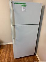 Front view of the white GE refrigerator showing the two doors and white handles, standing on a wood floor by a wall.
