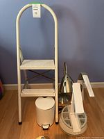 Photo showing white metal 2-step ladder, metallic desk light with cone-shaped shade, white desk light with round storage compartments base, white rectangular desk light, and small white garbage can.