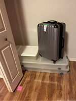 Three plastic storage containers stacked with large silver hard-shell suitcase on top, showing closed containers in a basement room with wood floor and white door partially visible.