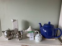 Photo showing full set of stainless steel and ceramic teapots, milk container, and other items on shelf.