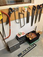 Photo of various hand saws hanging on a wall, a metal toolbox, black basket with extension cords, black plastic organizer box with nails and screws, and a spirit level on the floor.