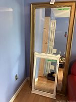 Shows two framed wall mirrors leaning against a blue wall: a large gold-colored framed mirror and a smaller white painted framed mirror.