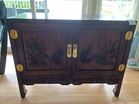 Front view of wooden cabinet showing carved bird and grass designs on double doors with brass hardware.