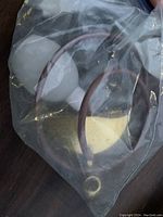 Brass powder sprayer enclosed in a clear plastic bag laying on a dark wood surface, showing its brass body and curved nozzle with flexible rubber tubing.