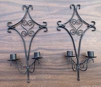 Pair of wrought iron wall hanging candle holders with symmetrical diamond design and two candle cups each, displayed on wooden background.