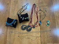 Full view of assorted costume jewellery on wood surface including necklaces, ankle bracelets, and earrings, displayed partially on two black fabric display pillows.