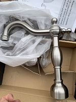 Brushed nickel finished bathroom faucet showing curved spout and ornate handle design, displayed partially wrapped in protective material inside a cardboard box.