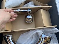 Hand holding a polished chrome lever handle with round base and curved spout lying in a box with packaging materials.