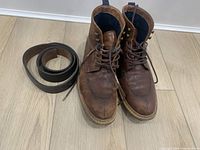 Pair of brown men's leather boots and a rolled-up leather belt placed on a wooden floor.