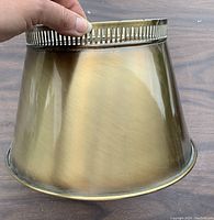 Metal brass lamp shade held by hand, showing exterior perforated edge and conical form