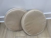 Two beige velvet round cushions resting on a wooden floor against a white wall, showing shape and fabric texture.