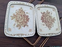Pair of foldable tables with metal floral trays on a wooden surface. Both trays have cream background with brown and beige floral bouquets and decorative border patterns.