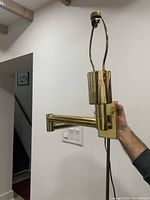 Photo of a gold finish wall-mount swing-arm light held by hand showing side and front with visible power cord and mounting plate