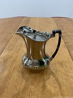 Front and side views of a small vintage metal pitcher with a black tubular handle that shows signs of repair on a wooden surface.