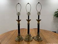 Front view of three brass and black finish stick table lamps on a wooden table, showing harp frames and lamp sockets minus shades.