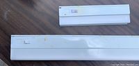 Photo showing two white under cabinet light fixtures side by side with on/off switches visible. Visible surface wear on one light.