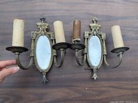 Pair of vintage two-light wall sconces shown side-by-side. Both have oval beveled mirrors, metal decorative frames, and candle-style holders with sleeve covers.