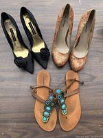 Top view of three pairs of shoes on wooden floor: black high heels with flower detail, floral pattern high heels, tan flat sandals with blue and green beaded decorations.