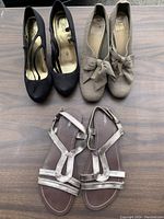 Three pairs of women's shoes arranged on a wooden surface: black heels with straps, grey flats with bows, and silver flat sandals with crisscross straps.