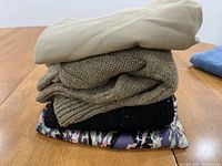 Stack of folded clothes showing top beige skirt, olive green knitted sweater, black knitted sweater, and camouflage-patterned fabric at bottom.