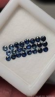 24 loose blue sapphires arranged neatly in a white display case, clear view of the stones' size and cut.