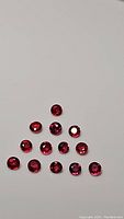 Twelve round calibrated rubies arranged in a triangular formation on a white background showing deep red color with pink-purple undertones and high clarity.