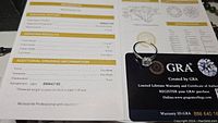 Photo showing GRA certification report with grading results: 1.0 carat, D color, VVS1 clarity, excellent cut, polish and symmetry. Moissanite ring visible on report and certification card.
