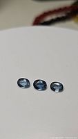 Three oval cut denim blue sapphires placed on a white surface showing the stones' color and clarity.