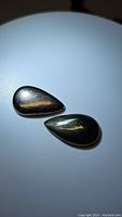 Pair of pear-shaped golden pyrite cabochon stones with smooth polished surfaces and metallic shine placed on a white background.