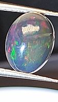 Close-up photo of oval cabochon Ethiopian black opal held with tweezers showing rainbow color flashes on black base.
