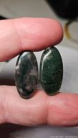 Two oval-shaped moss agate cabochons held between fingers, showing distinct moss-like internal patterns and polished oval surfaces.