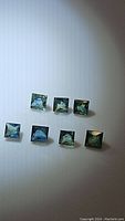 Seven multicolored princess cut sapphires arranged on white surface in two rows, showing their green to blue colors and clarity.