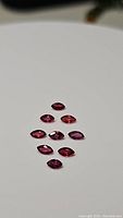 Nine marquis cut sapphires in shades of red to pink arranged on a smooth white surface, showing their faceted cuts and colors.