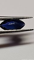 Close-up photo of a deep blue marquis cut sapphire held by tweezers, showing color and cut details.