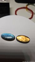 Pair of natural labradorite oval cabochons placed side by side on a light surface, one displaying blue flash and the other golden flash.