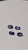 Four oval cut natural tanzanite gemstones laid on a plain white background, showing blue-purple color and oval faceted shape.