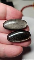 Two oval cabochons of silver sheen obsidian held on fingertips showing metallic silver flash.
