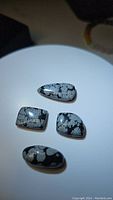 Photo showing four snowflake obsidian cabochon gemstones laid out on a white round base. The stones possess different shapes and exhibit characteristic white snowflake patterns on a black background.