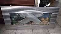 Front view of rectangular in-wall style aquarium with brushed metal wavy frame, showing gravel and decorative interior elements.