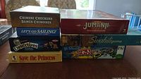 Seven board game boxes stacked: Chinese Checkers, Jumanji, Let's Go Sailing, We Detectives, Swahili game, Save the Princess, Spin Pilot.