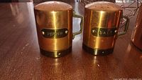 Close-up of brass salt and pepper shakers showing labels and perforated tops, placed on wood surface.