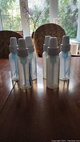 Six clear plastic Dr. Brown's baby bottles with white caps on a wooden table, showing full bottles with internal blue vents and caps attached.