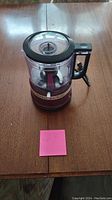 Small KitchenAid food processor with clear plastic bowl and lid, black handle on maroon base.