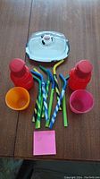Photo of two red kids water bottles, two small colorful cups (orange and pink), nine assorted silicone straws, and transparent butter dish with cow design lid on wooden surface.