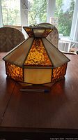 Hexagonal stained glass hanging light fixture with amber and cream colored glass panels on a wood table, showing overall shape and details.