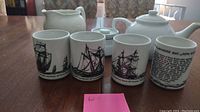 Photo showing four ceramic teacups featuring black ship illustrations and text, alongside sugar bowl, creamer, and teapot parts.