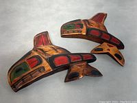 Two hand-carved cedar killer whale carvings with red, green, and black paint accents in traditional First Nations style, placed on a gray surface.