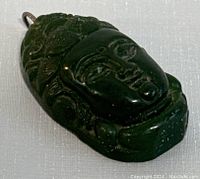 Close-up front view of deep green jade Buddha face pendant showing carved details and metal loop at top.